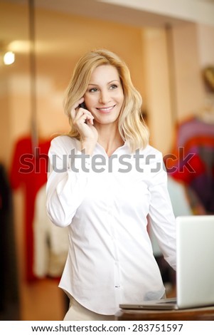 Portrait of busy small clothing store owner standing in front of laptop and making call. Small business.
