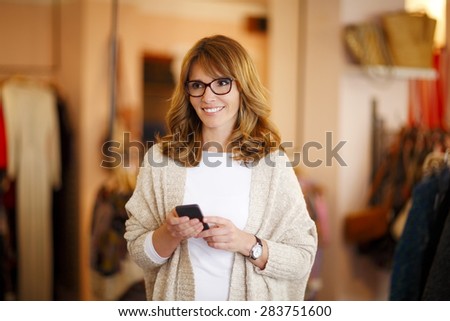 Portrait of middle age businesswoman holding smart phone and texting message while standing in her small store. Small business.
