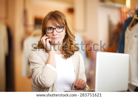 Portrait of small clothing store owner woman making call while standing in her small shop in front of computer. Small business.