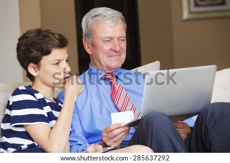 Portrait of grandfather with laptop and his grandson with digital tablet sitting at home and using credit card for online shopping.
