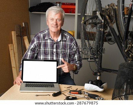 Portrait of senior bike shop owner sitting at desk behind the laptop and points to the white screen of the laptop.
