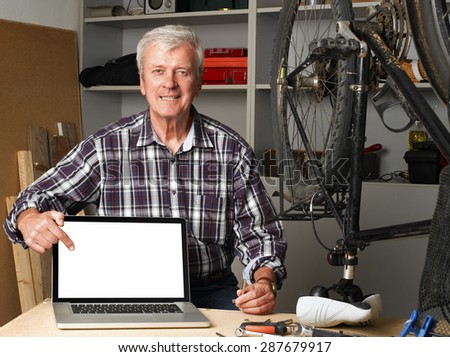 Portrait of senior bike shop owner sitting at desk behind the laptop and points to the white screen of the laptop.