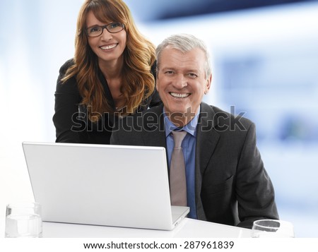 Portrait of happy senior businessman sitting behind the laptop while sales woman standing next to colleague and looking at camera. Teamwork at office.
