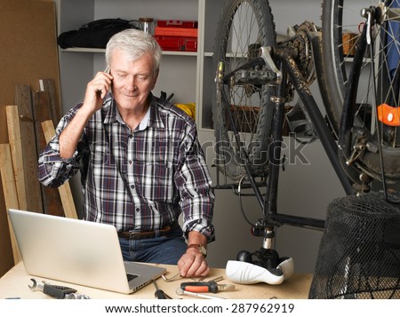 Portrait of senior man sitting at his bike shop and repairing bicycle while making call. Small business.