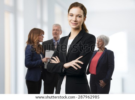 Portrait of young businesswoman standing at office with arms crossed while her colleagues consulting at background.