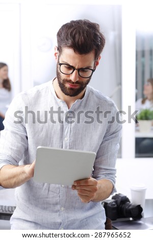 Portrait of young handsome sales man sitting at office desk in front of computer while holding digital tablet and touch the screen.