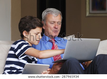Portrait of young teenager boy with digital tablet sitting at sofa at home with his grandfather and teaching how to use the laptop. Grandson points out the screen of the laptop.