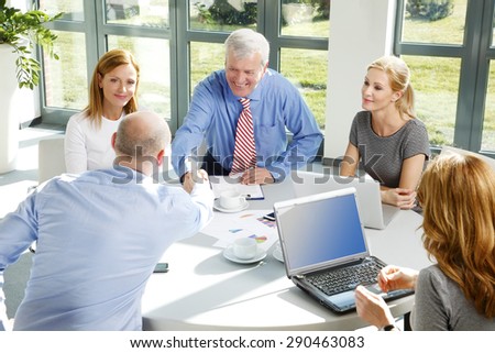 Portrait of two businessmen shaking hands while making agreement and sitting at business meeting. Business people using laptop and digital tablet while sitting around conference table.