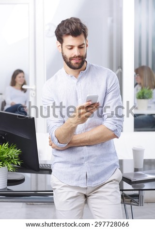 Portrait of young professional standing at office while using his mobile. Creative manager checking emails on his handy.