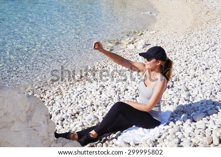 Full length portrait of fit woman sitting at the pebble beach after morning run and taking self portrait with mobile phone.