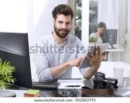 Portrait of young newspaper editor sitting at workplace and holding his hands digital tablet while working online.