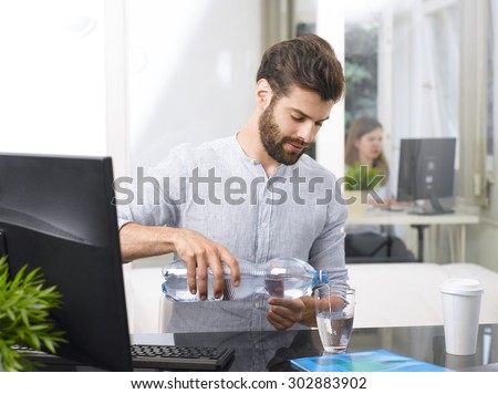 Portrait of young businessman sitting at his working place and pouring water into a glass.