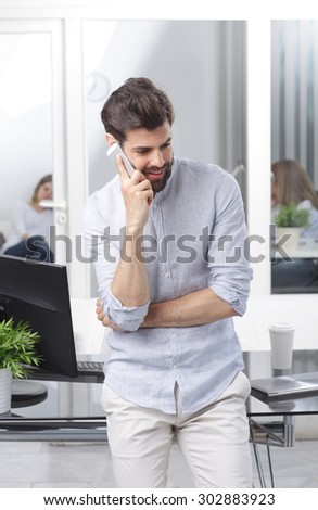 Portrait of young professional man standing at desk in front of computer while making phone conversation.
