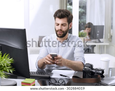 Portrait of young creative sitting at office in front of computer and writing email on his mobile. Small business.