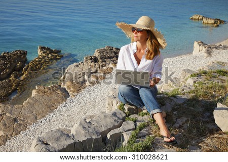 Portrait of smiling woman working with laptop while sitting on the beach on vacation.