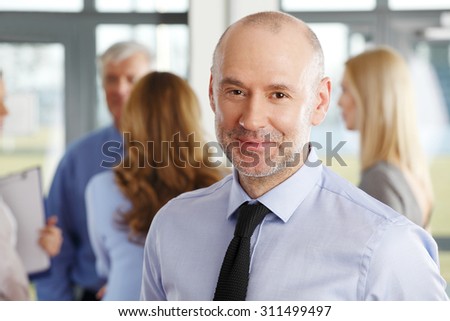 Close-up portrait of senior businessman standing at business meeting while looking at camera and smiling. Business people standing in background.