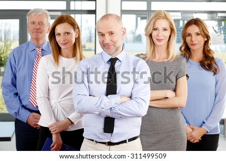 Portrait of successful sales man with arms crossed standing at office with his sales team. Mixed age businesswomen and businessmen in row.
