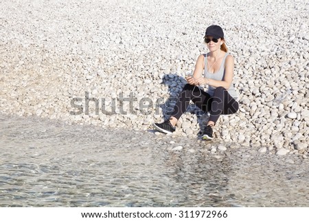 Full length portrait of sporty woman sitting on the beach and relaxing after morning running. Sporty female listening