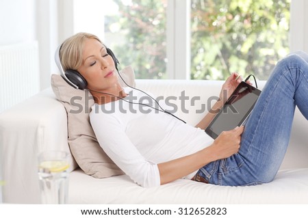 Portrait of beautiful woman relaxing at home. Beautiful female lying on sofa while holding hand digital tablet and listening music with headphone.