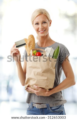 Portrait of smiling woman standing at supermarket and holding hand shopping paper bags full of food while other hand there is a credit card.