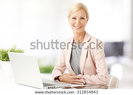 Portrait of middle age woman sitting at office and working. Beautiful businesswoman sitting at desk in front of laptop while looking at camera and smiling.