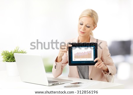 Portrait of middle age businesswoman holding hand touchpad with column cart on the screen while sitting at office and analyzing financial data.
