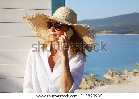 Close-up portrait of smiling woman standing by the beach house and making call during her summer vacation.