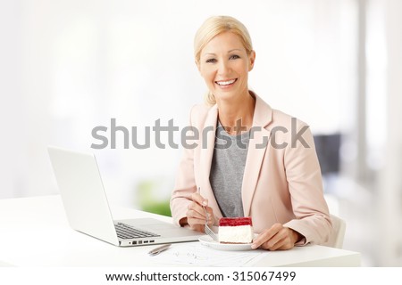 Portrait of smiling businesswoman sitting at office in front of laptop and eating a piece of cake while working.