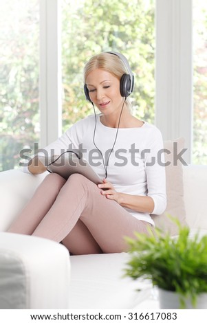 Portrait of beautiful middle age woman listening music while sitting at home on the sofa. Smiling female with headphone using digital tablet.