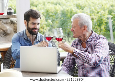 Portrait of young and senior man tasting with red wine in a vineyard. Professional winemakers sitting at desk in front of laptop at wine cellar and consulting. Multi generation small business.