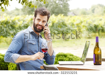 Portrait of young winemaker sitting at wine cellar in front of his laptop. Smiling professional man holding in her hand a glass of red wine and tasting it while making call. Small business.