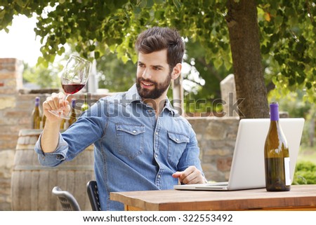 Portrait of young winemaker holding in his hand a glass of red wine and tasting. Professional man sitting at wine cellar in vineyard and working on laptop. Small business.