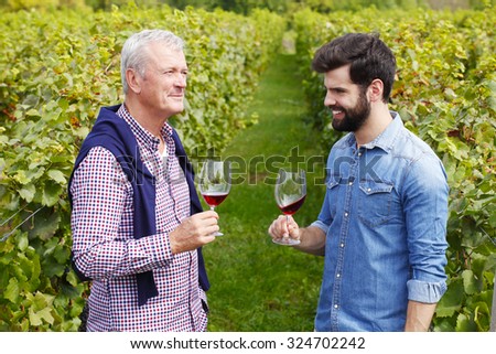 Portrait of multi-generation family working together. Senior winemaker and young professional man standing at vineyard while holding in their hands a glass of wine and tasting it. Small business.