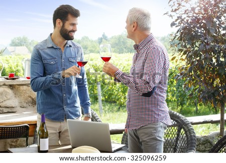 Portrait of young winemaker and senior sommelier toast with red wine while standing at family wine cellar and working with laptop.