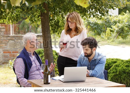 Multigenerational family working together. Middle age woman holding a glass of red wine and tasting it. Senior sommelier and young professional man working on laptop at wine cellar and consulting.