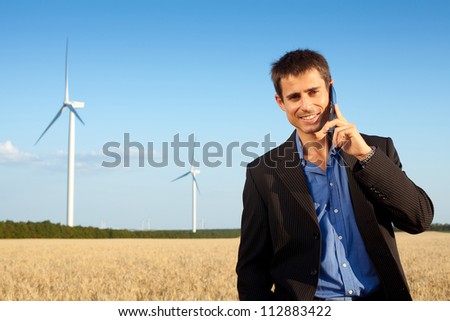 smiling farmer (businessman) standing in wheat field over wind turbines background and speaking on mobile phone