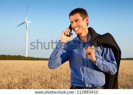 smiling farmer (businessman) standing in wheat field over wind turbines background and holding his mobile phone and jacket over shoulder