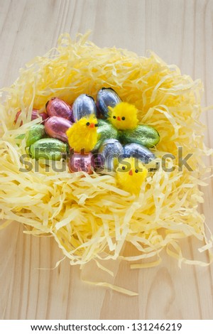 Easter chocolate eggs, wrapped in shiny colorful foil, placed in a nest made of yellow, shredded tissue paper looking like  hay. Little chicken toys also sit in the nest.