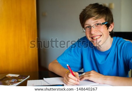 Smiling teenager studying and doing his homework while holding his pen and writing in his notebook.