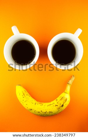 Two coffee cups and a banana shot on an bright orange background. The composition of the objects looks like a smiley face.