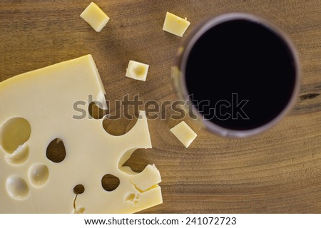 A block of cheese on a wooden cutting board shot from above. Some of the cheese is cut into small cubes. A glass of red wine is placed alongside.