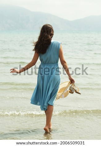 Back view of a young brunette woman in a blue dress walking barefoot on a beach and dangles his feet in the water. Foto d'archivio © 
