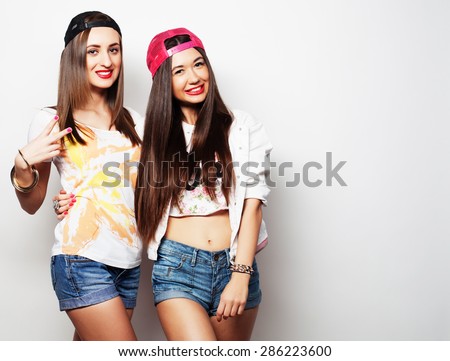 Two young girl friends standing together and  looking at camera. Over grey background.