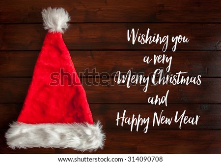 Red santa hat with white fur trim on brown wooden floorboards with Christmas greeting text beside it.