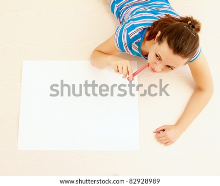 Top view of cute young woman thinking with a pen in her mouth while lying on floor and drawing.
