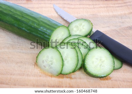 A fresh green English cucumber cut into lots of slices with a knife on a wooden board.