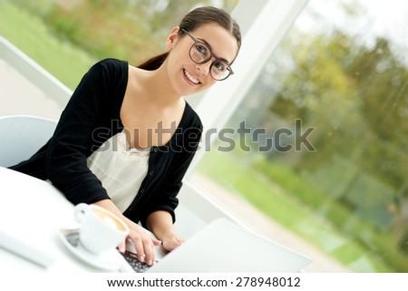 Smiling young woman relaxing with her laptop computer at home sitting at a table on her outdoor patio wearing sunglasses with a cup of fresh hot coffee alongside