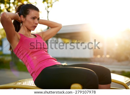 Side view of woman sitting on a bench outside doing situps with hands behind head.
