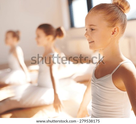 Close up side view of the face of a cute pretty little blond ballerina smiling in class as she practices her poses with her classmates in a warm bright ballet studio