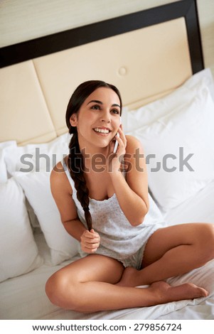 Pretty young woman with her long hair in a braid talking on her mobile phone as she sits on top of her bed in her sleepwear enjoying a relaxing morning